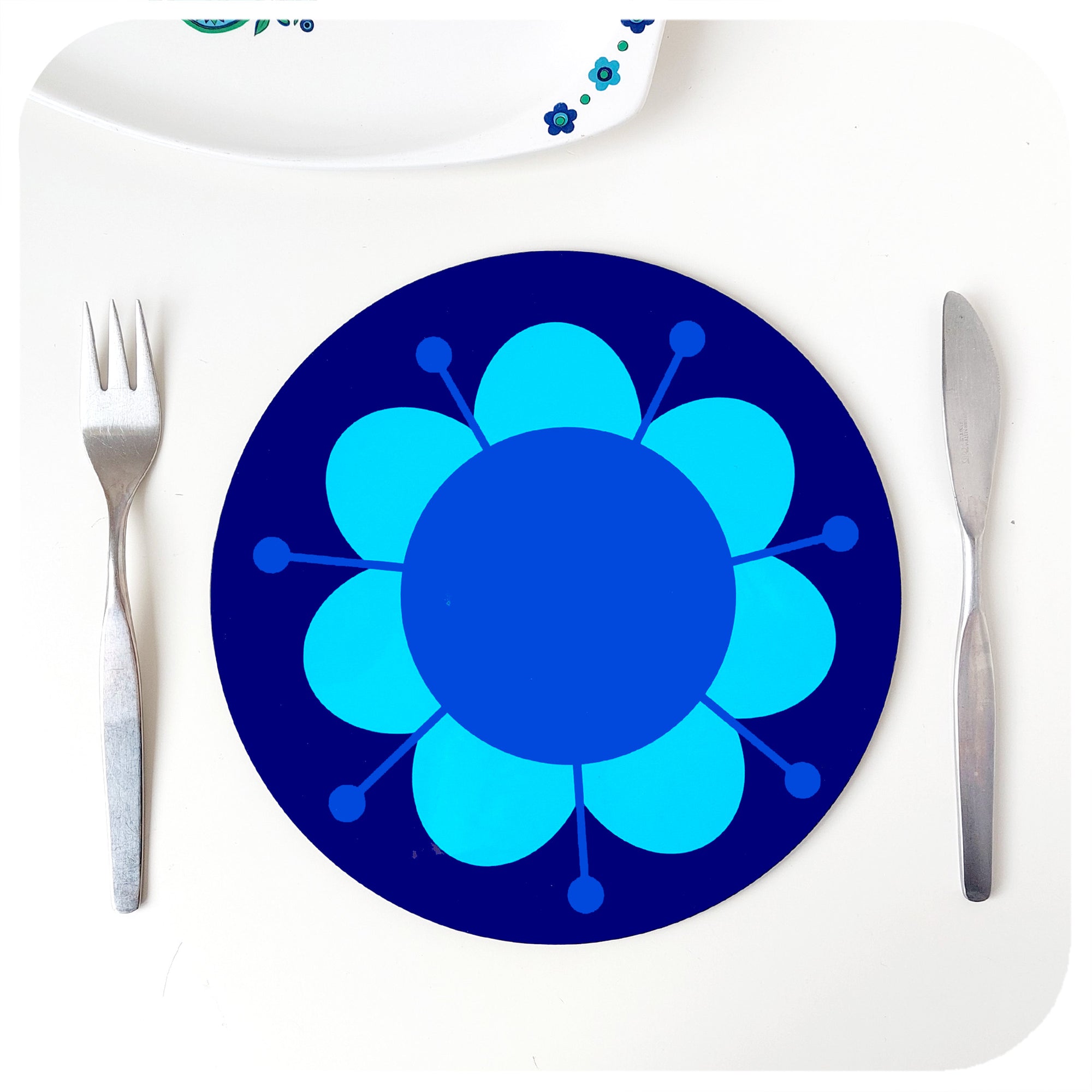 A round retro floral placemat featuring our Daisy design, in blues & purples, on a white table with cutlery and vintage serving dish | The Inkabilly Emporium