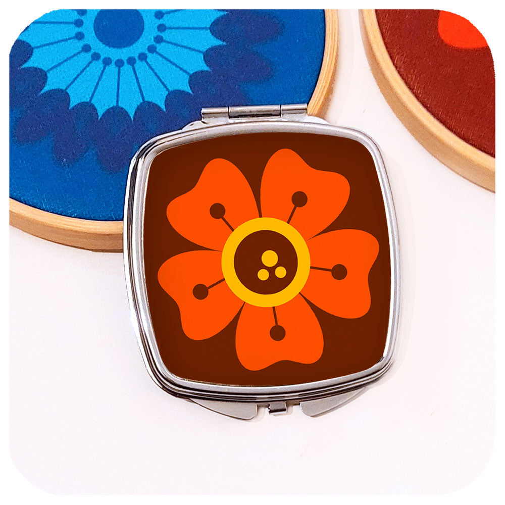 A chrome compact mirror with a 70s style retro flower design on the front leans against a bamboo hoop with fabric in similar colours, on a white background | The Inkabilly Emporium