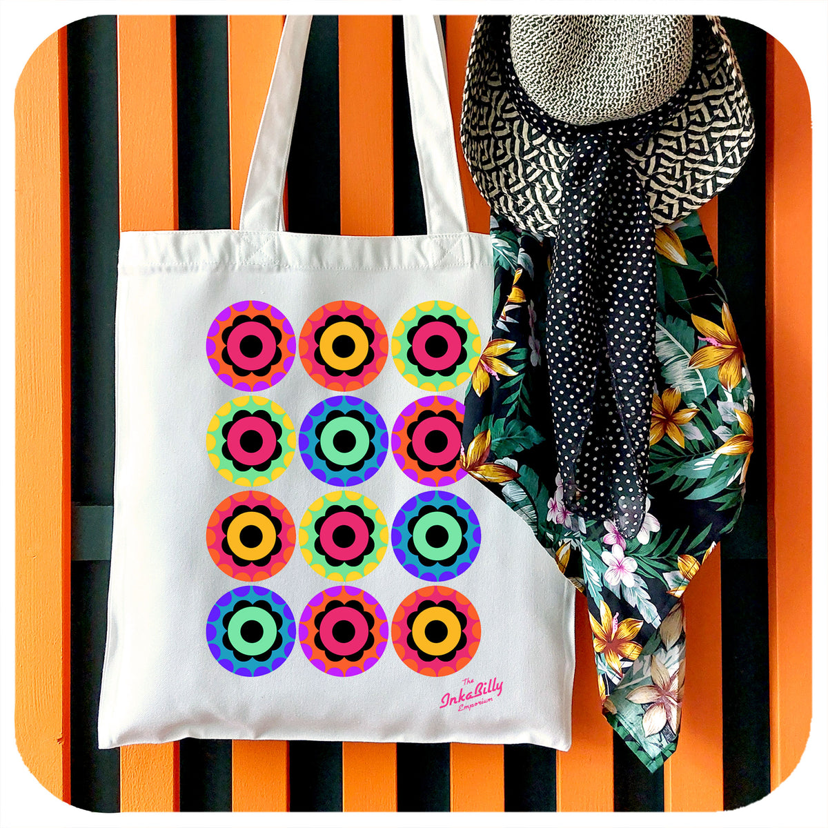 A canvas style tote bag with a 60s style flower power print hangs on a coat rack with a straw sun hat and tropical print bandana | The Inkabilly Emporium
