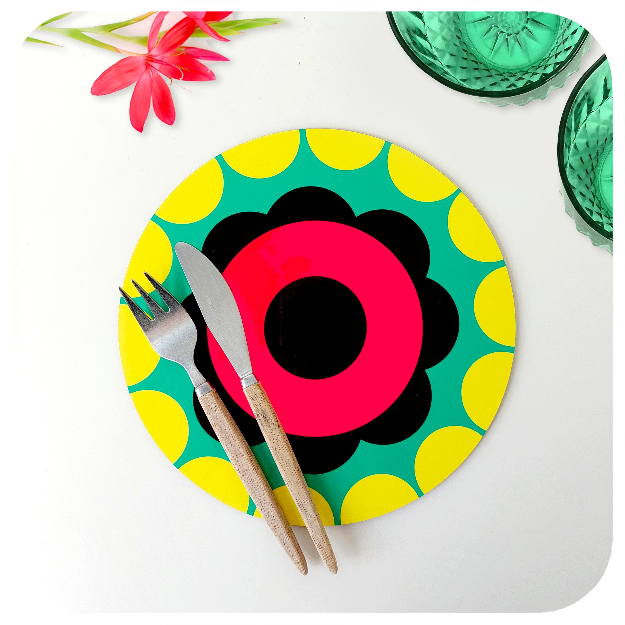 A multi-coloured 60s style flower placemat in pink, green & yellow,  is set on a white table with cutlery, green glass bowls and a pink flower | The Inkabilly Emporium