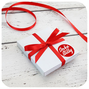 A small, white, square cardboard gift box, is tied with red ribbon and has an Inkabilly logo sticker. It is lying on a white wooden surface next to a piece of loose red ribbon | The Inkabilly Emporium