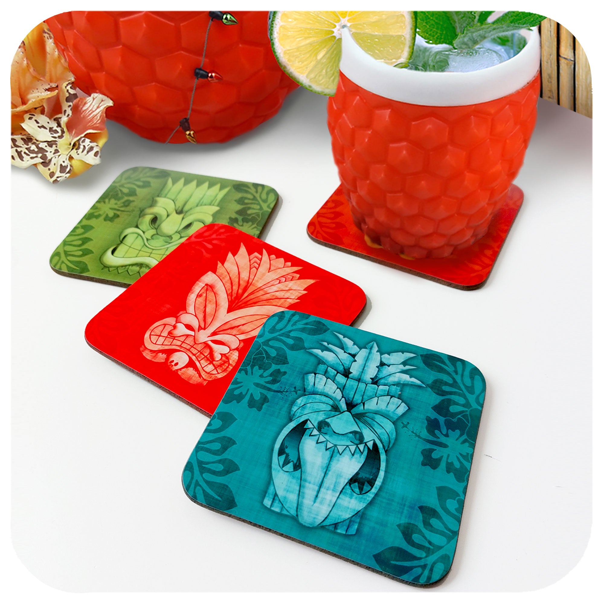 Set of four Tiki coasters in various colours sit on a white table with an orange plastic tumbler containing a cocktail, plus other tropical accessories in the background | The Inkabilly Emporium