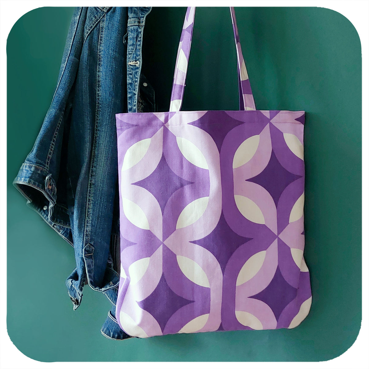 A handmade tote bag, made from vintage 70s fabric, with an Op Art pattern in purple & white, hangs against a green wall next to denim jacket | The Inkabilly Emporium
