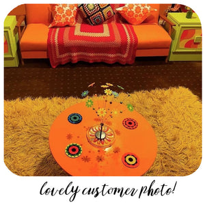 Customer photo of flower power style coasters sitting on a round, orange coffee table in a 60s style living room with shag pile rug, orange sofa with crochet cushions & blankets. Text along the bottom of the image reads : Lovely customer photo! | The Inkabilly Emporium