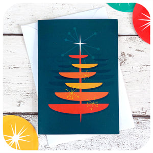 Mid Century style Christmas Card featuring an MCM style orange Christmas Tree on a blue background, with a white envelope, on a white table with 50s style xmas decorations | The Inkabilly Emporium