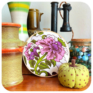 Vintage fabric with floral design, framed in an embroidery hoop sits with ceramic pots, and a small pumpkin on a wooden surface.