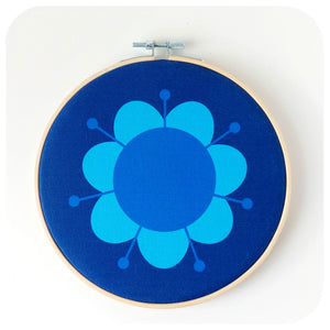 A round blue fabric wall art hoop featuring a hand-printed retro daisy design, framed in a bamboo embroidery hoop.