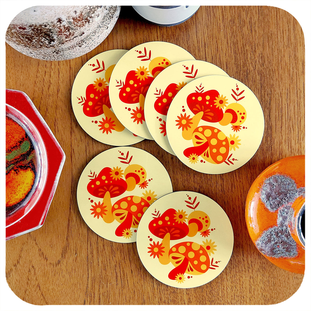 Set of 6 70s style retro coasters featuring orange, red & yellow mushrooms. They are lying on a teak table with various vintage pottery pieces around the edge of the frame. | The Inkabilly Emporium