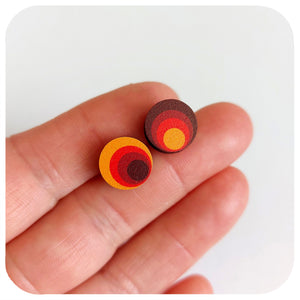 Round wooden earrings with concentric colour rings held between fingers on a white background