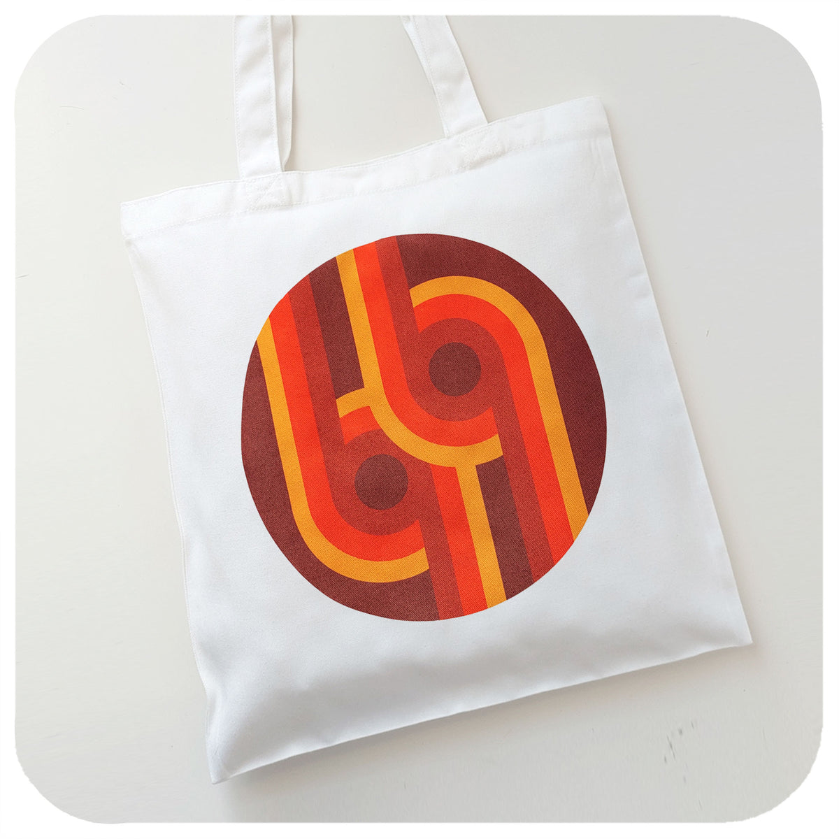 A canvas style tote bag featuring a 70s supergaphic print in orange, yellow, red and brown lies on a white background | The Inkabilly Emporium