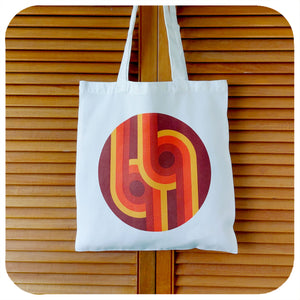 A canvas style tote bag featuring a 70s supergaphic print in orange, yellow, red and brown hangs against a pair of wooden louvre doors  | The Inkabilly Emporium