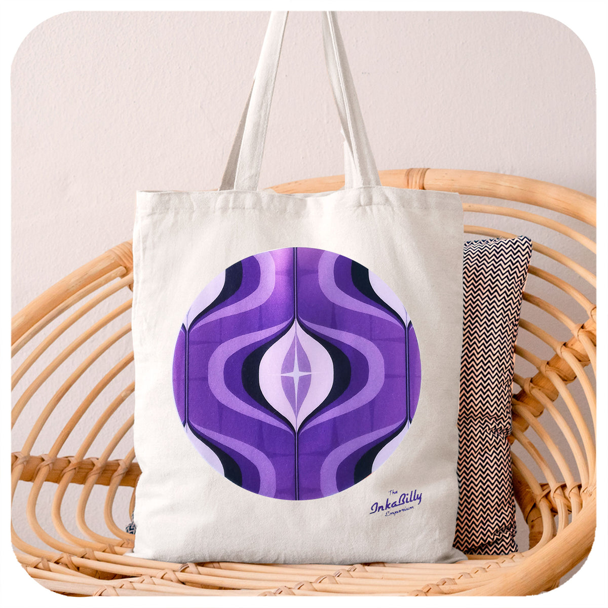 A white tote bag with a purple 70s style graphic, sits on a rattan chair | The Inkabilly Emporium
