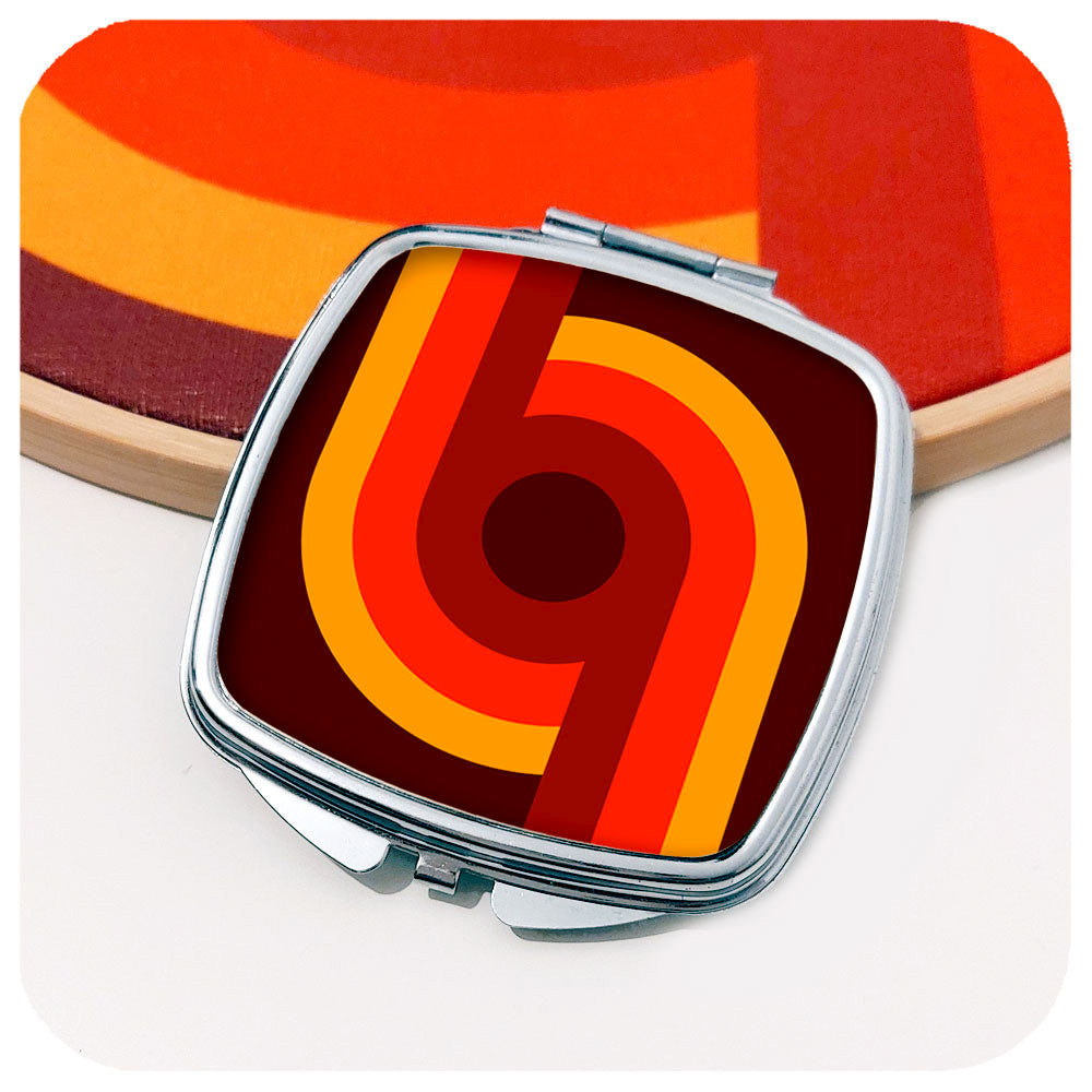 A chrome compact mirror with a 70s style graphic design on the front leans against a bamboo hoop with fabric in similar colours, on a white background | The Inkabilly Emporium