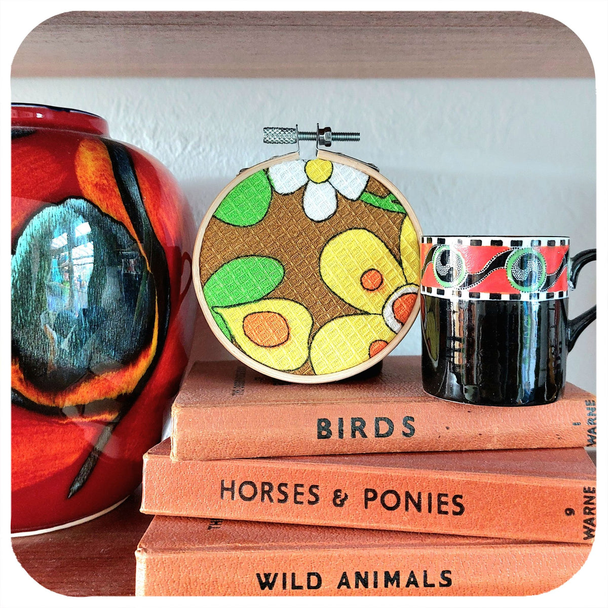 Colourful floral fabric framed in an embroidery hoop sits on a stack of books on a shelf next to vintage ceramics