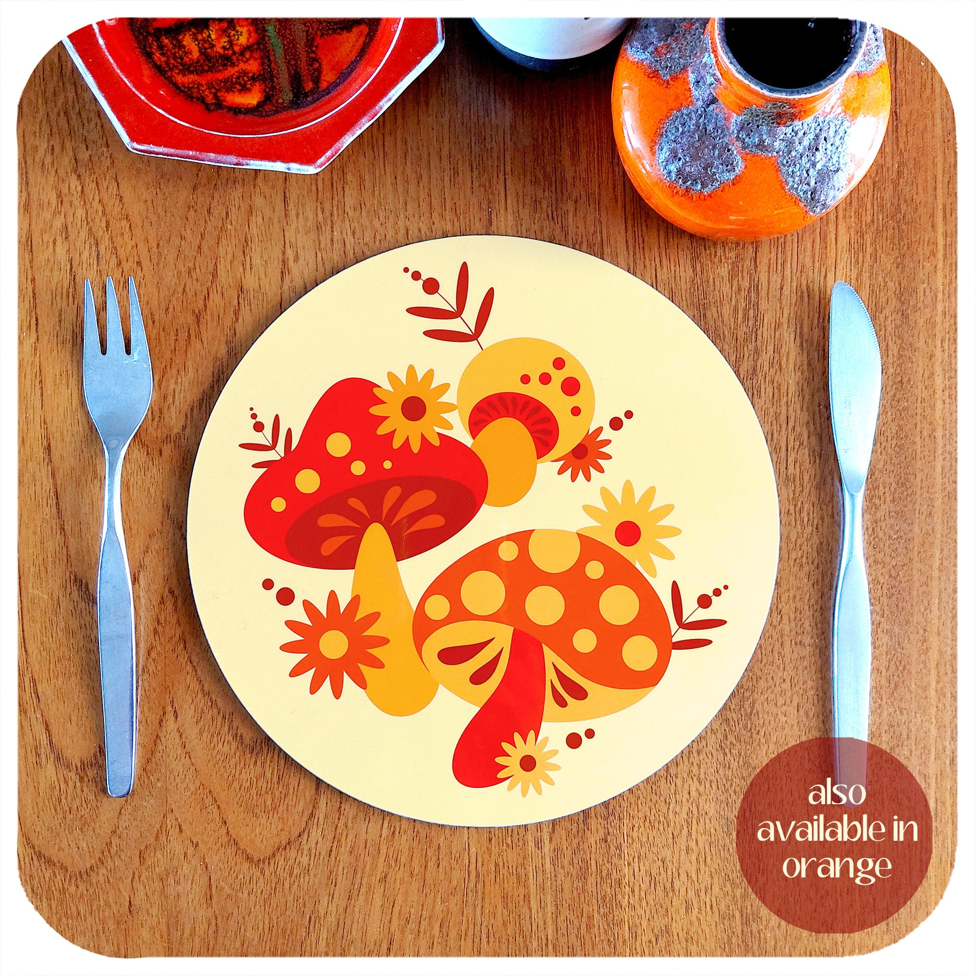 A round, 70s style placemat featuring retro mushrooms sits on a teak table with cutlery and vintage pottery pieces. Text in lower righthand corner reads: also available in orange | The Inkabilly Emporium
