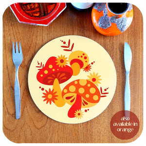 A round, 70s style placemat featuring retro mushrooms sits on a teak table with cutlery and vintage pottery pieces. Text in lower righthand corner reads: also available in orange | The Inkabilly Emporium