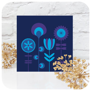 A square card featuring blue & purple scandi style flowers stands in front of a white brick wall with dried allium flowers | The Inkabilly Emporium