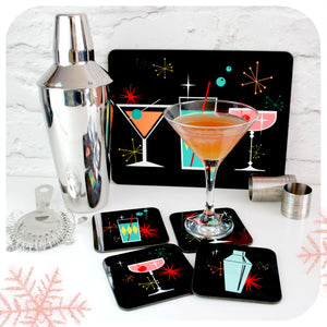 A placemat featuring mid century style cocktails leans against a white wall next to a cocktail shaker and martini cocktail, with matching coasters | The Inkabilly Emporium