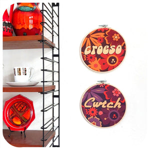 Decorative items including a shelf with red and orange vases, a white mug, and a red plate. Two embroidery hoops with floral patterns and text on a white background.