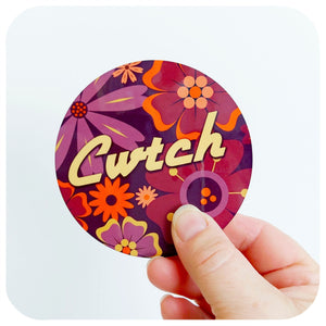 Colourful floral design coaster with 'Cwtch' text held by a hand on a white background