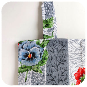 Close up of a floral patterned fabric tote bag with blue pansy and green leaves on a white background