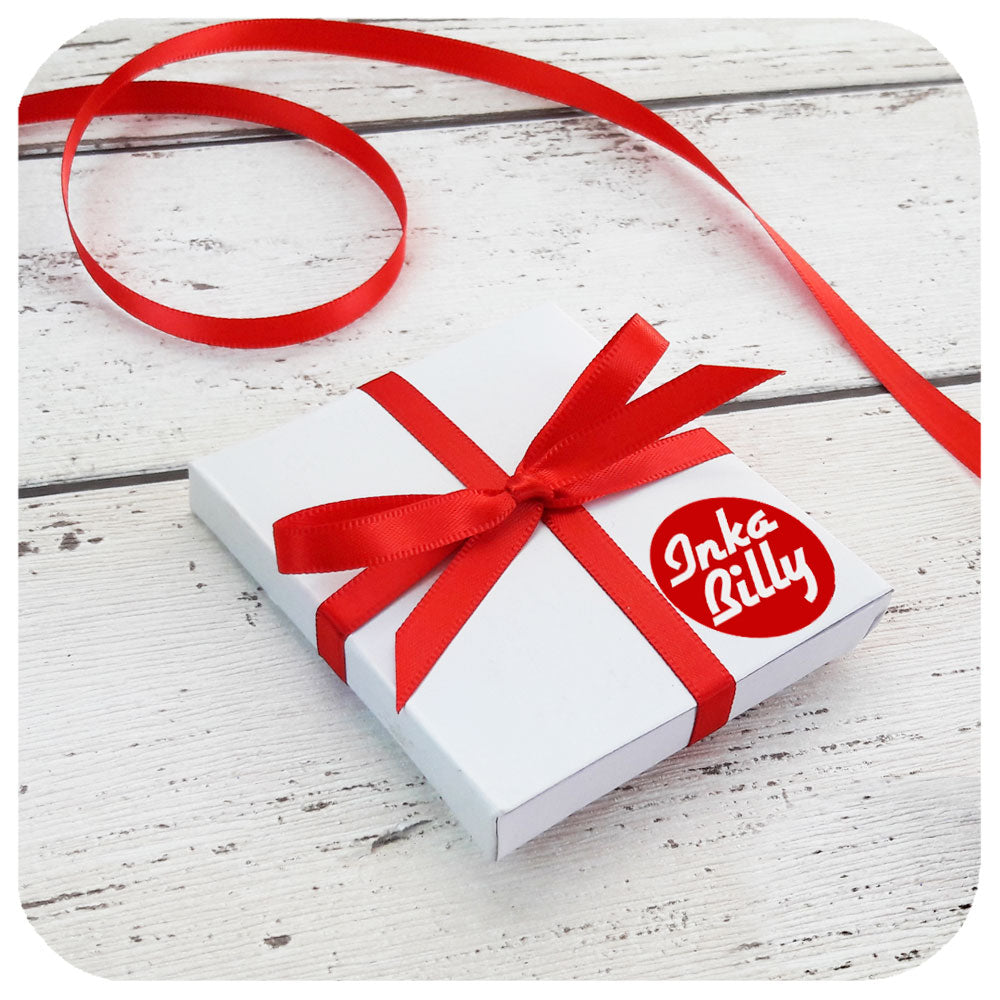 A square, white gift box tied with red ribbon and Inkabilly logo sticker, containing a compact mirror, on a white wooden background | The Inkabilly Emporium