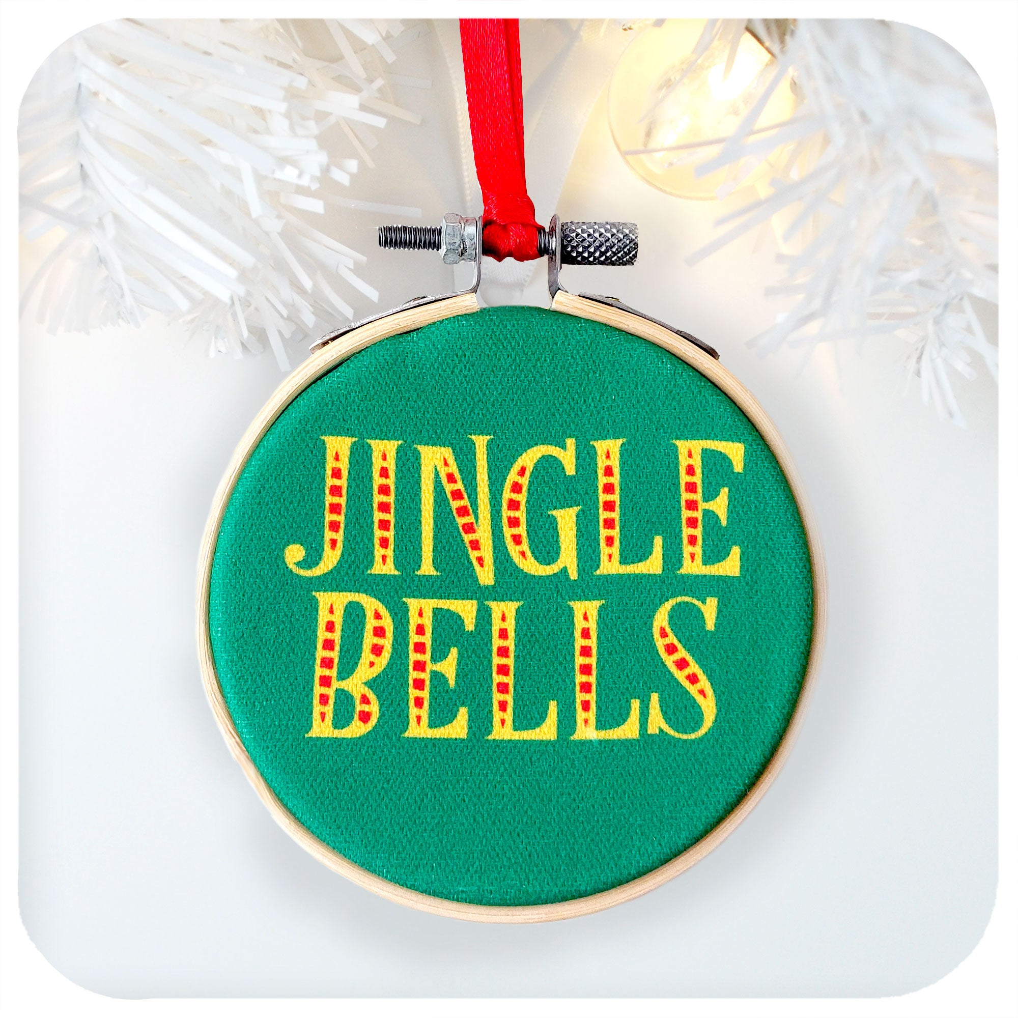 A printed fabric Christmas tree decoration on a white background with white christmas tree branch and fairy light. Text on the ornament reads: Jingle Bells | The Inkabilly Emporium