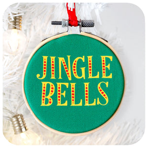 A printed fabric Christmas tree decoration on a white background with white christmas tree branch and fairy light. Text on the ornament reads: Jingle Bells | The Inkabilly Emporium