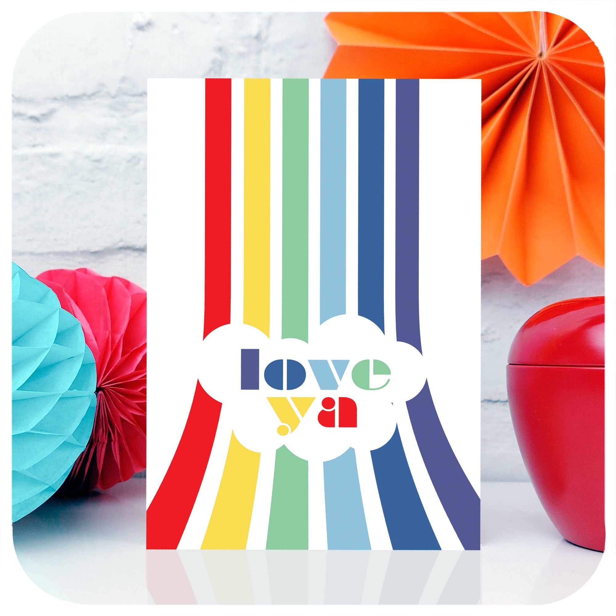 An A6 greetings card featuring a vertical rainbow with the words "Love Ya" stands on a white table with colourful party decorations | The Inkabilly Emporium