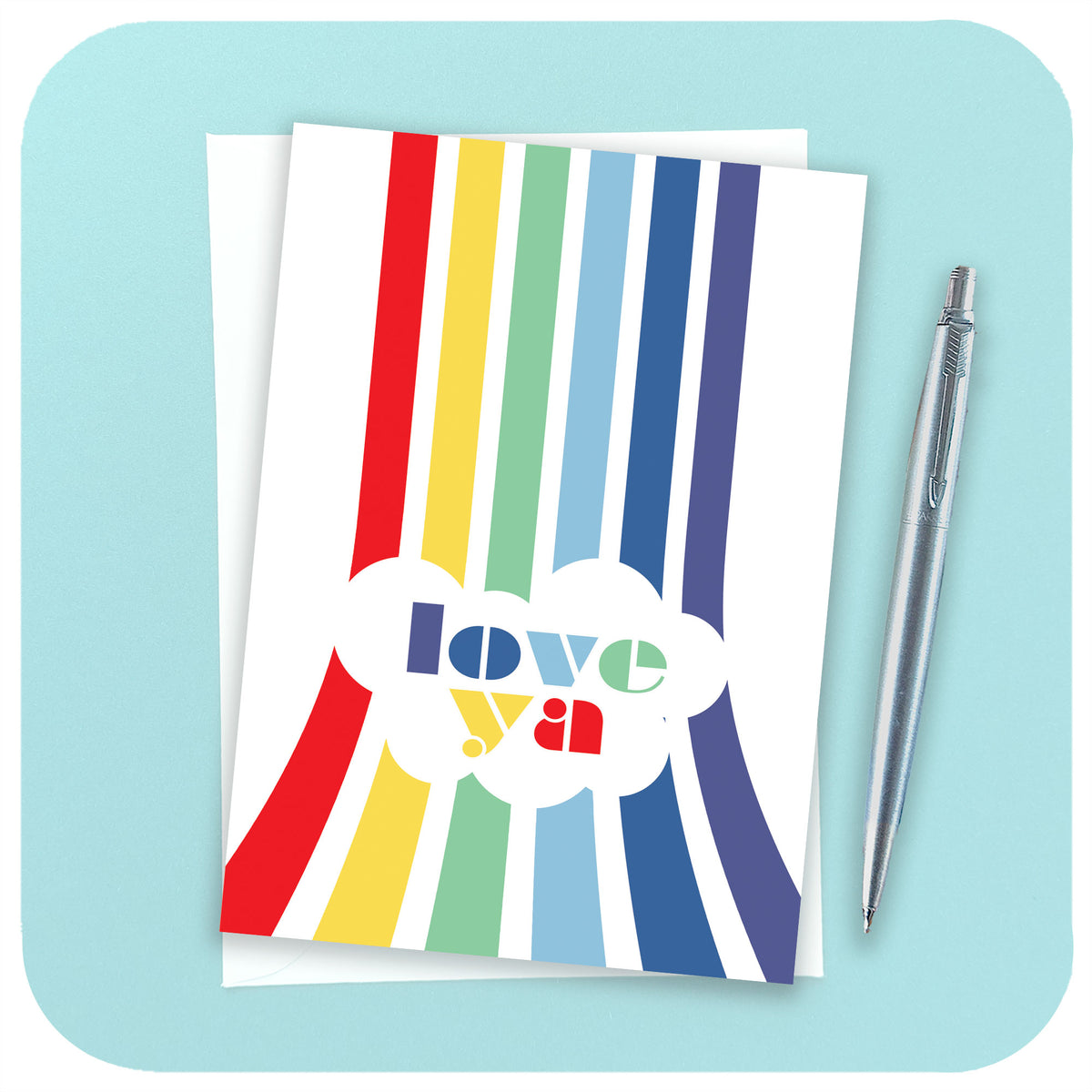 An A6 greetings card featuring a vertical rainbow with the words "Love Ya" sits on a white envelope beside a chrome pen on a light blue background | The Inkabilly Emporium