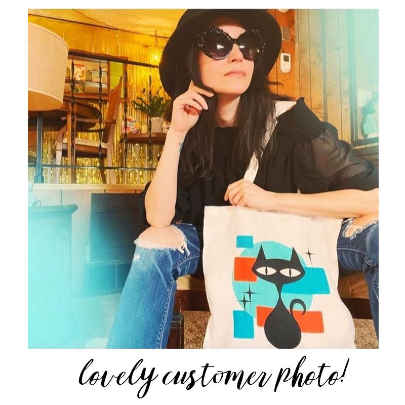 Customer photo of a white woman in big sunglasses and hat sitting, holding a tote bag featuring a reto atomic style cat. Text along the bottom of the image reads : Lovely Customer Photo! | The Inkabilly Emporium 
