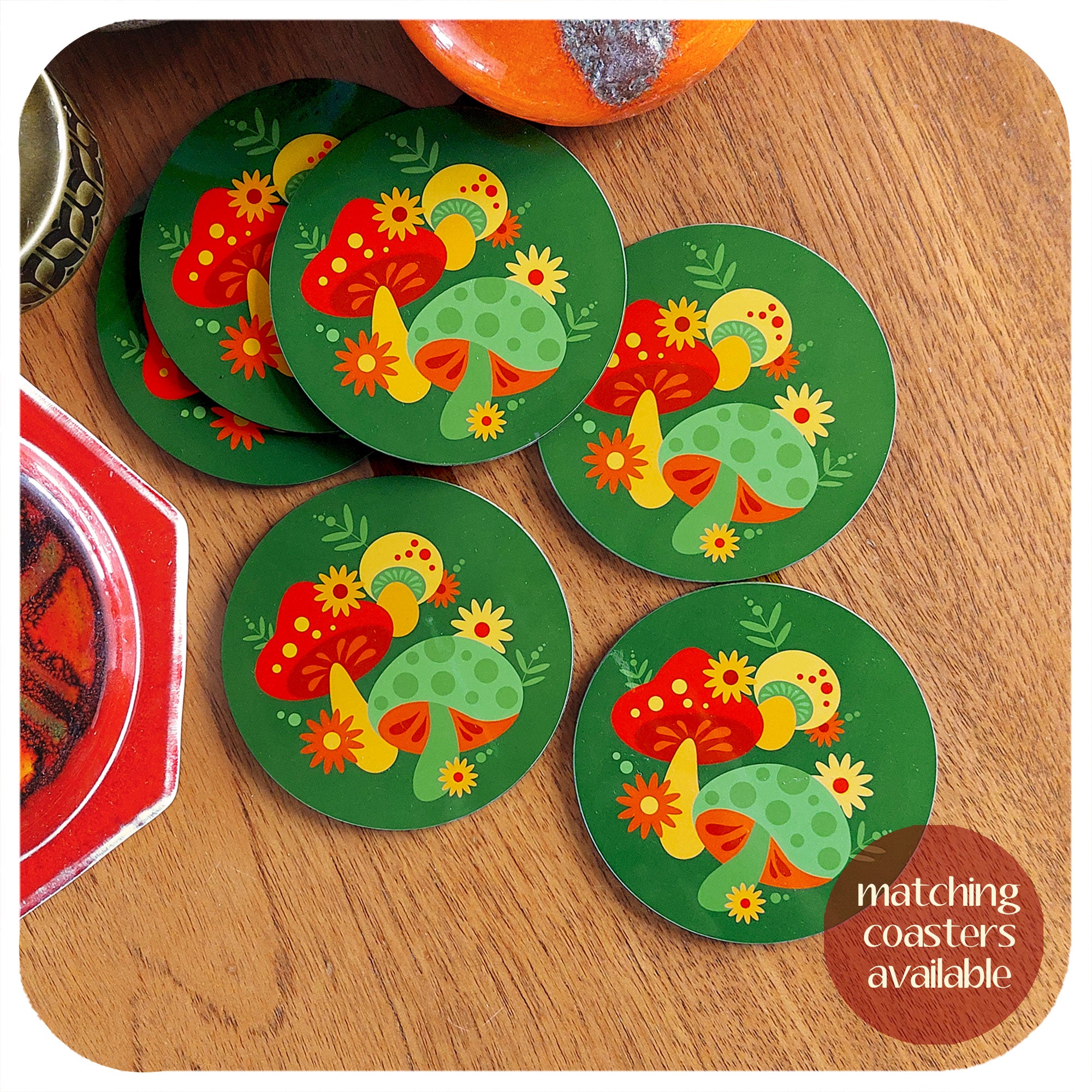 A set of six round vintage style mushroom coasters sit on a teak table with vintage pottery pieces in the background. Text in the corner reads "matching coasters available" | The Inkabilly Emporium