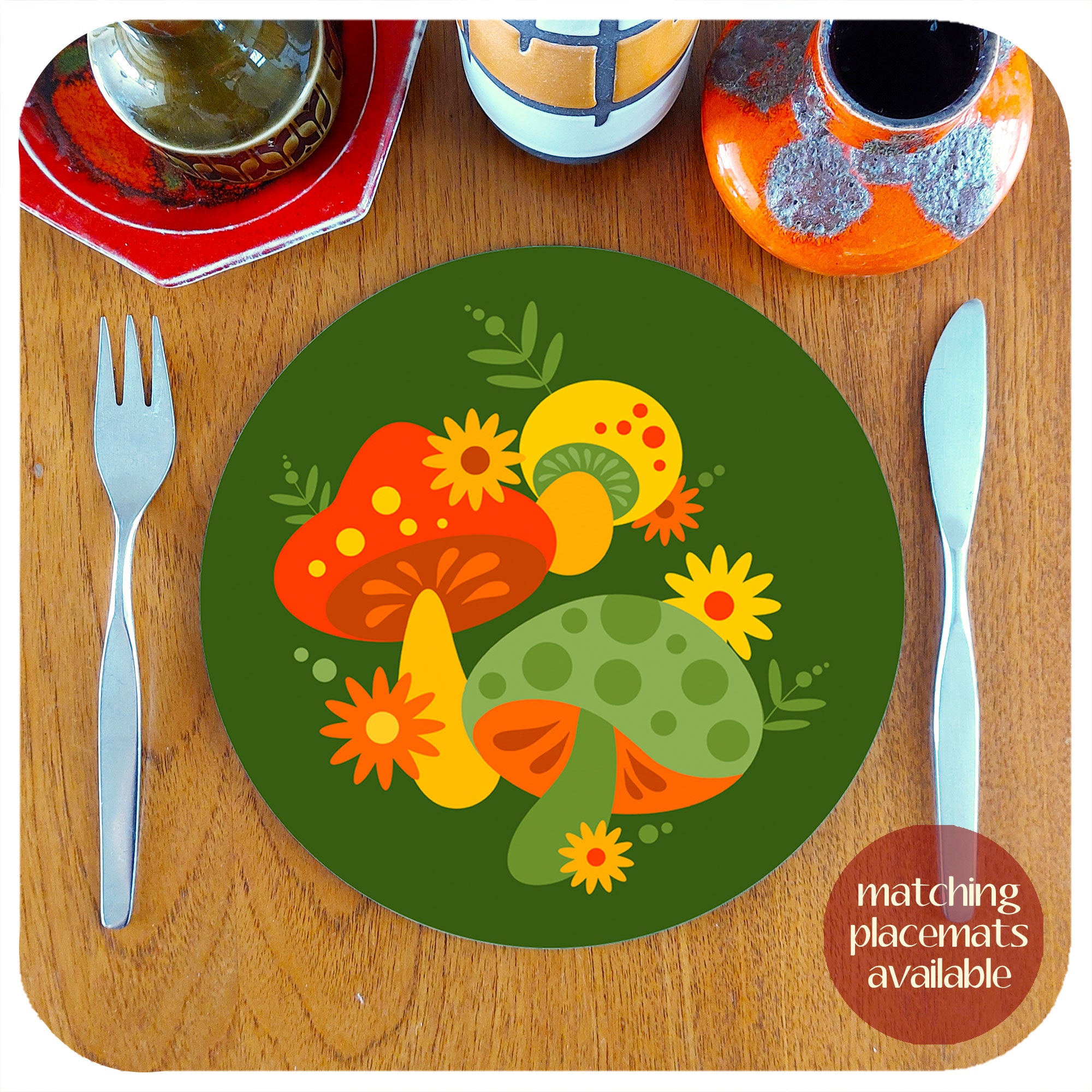 Round 70s style placemat featuring a retro mushrooms design sits on a teak table with mid century cutlery, accompanied by a selection of vintage pottery pieces. Text in the bottom right hand corner reads: matching placemats available | The Inkabilly Emporium