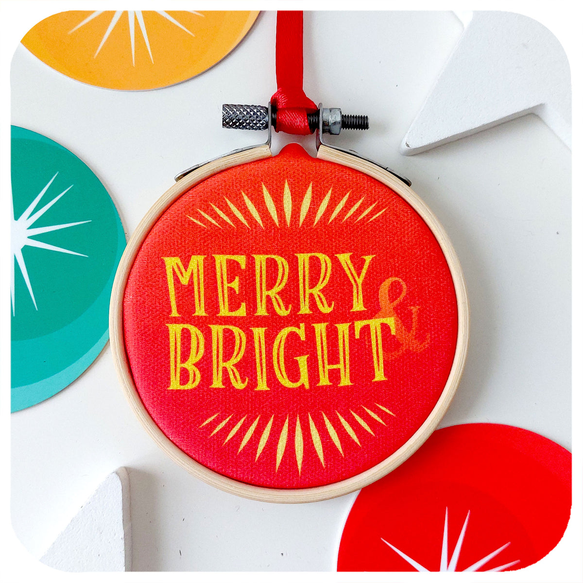 A printed fabric retro style Christmas tree decoration on a white background surrounded by colourful Christmas decorations. Text on bauble reads: Merry & Bright