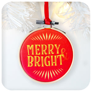 A printed fabric Christmas tree decoration on a white background with white christmas tree branch and fairy light. Text on the ornament reads: Merry & Bright
