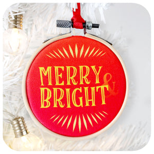 A printed fabric Christmas tree decoration hangs on a white Christmas tree. Text on the bauble reads: Merry & Bright