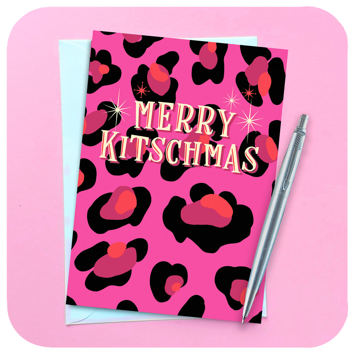 A PINK LEOPARD PRINT CHRISTMAS CARD LIES ON A PINK BACKGROUND WITH ENVELOPE AND PEN. TEXT ON THE CARD READS: MERRY KITSCHMAS.