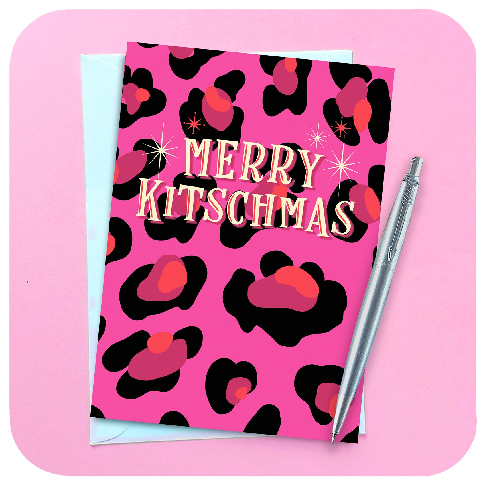 A pink leopard print christmas card lies on a pink background with a silver pen and white envelope. Text on the card reads: Merry Kitschmas.