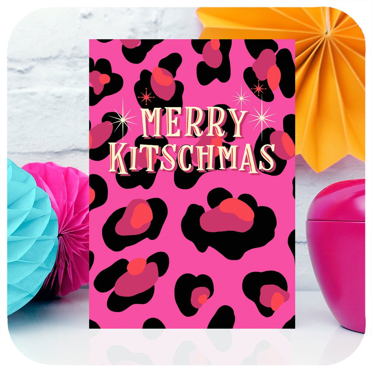 A pink leopard print christmas card stands among brightly coloured paper decorations. Text on the card reads: Merry Kitschmas.