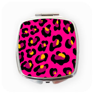 A square, chrome compact mirror with a pink leopard print design against a white background