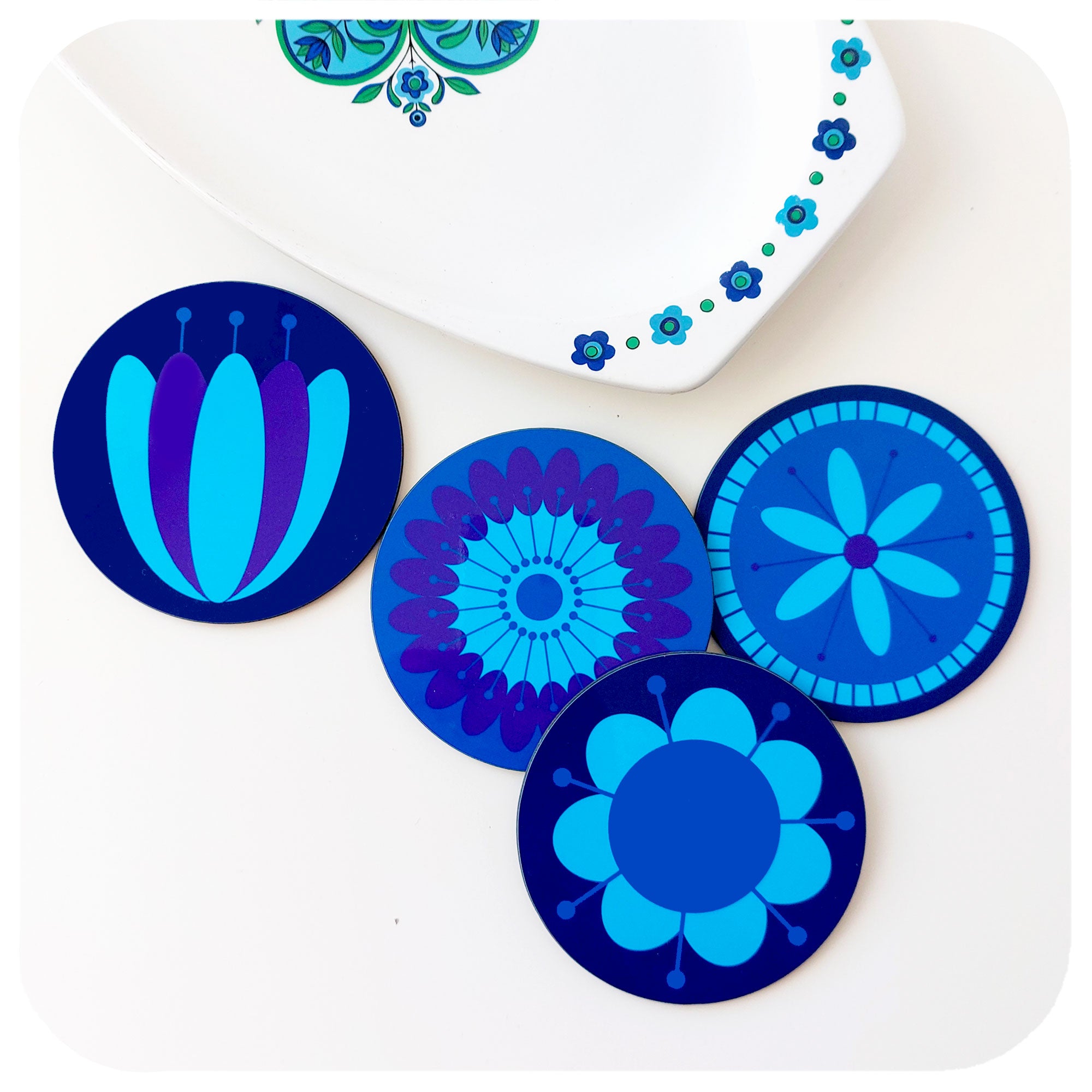 Four round coasters featuring retro style flowers in blues & purples sit next to a vintage J & G Meakin dish in similar colours on a white table | The Inkabilly Emporium