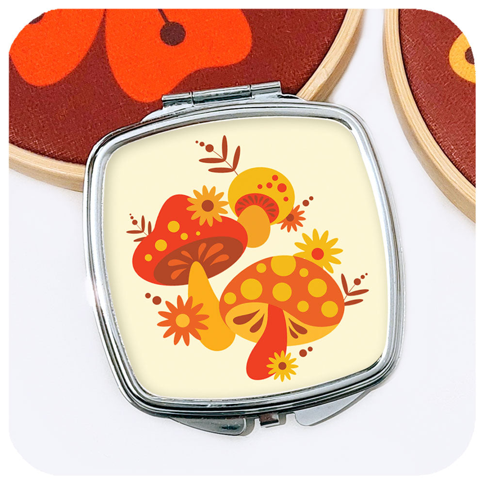 A square, chrome compact mirror with 70s style mushrooms motif leans against the edge of an embroidery hoop on a white table | The Inkabilly Emporium