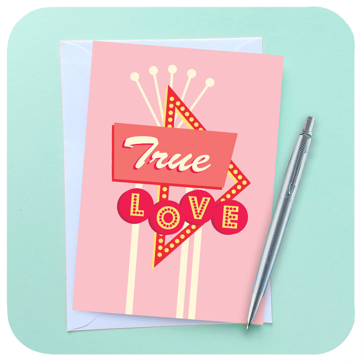 A pink A6 greetings card featuring a  graphic rendering of a 50s style American roadside neon sign with the words "True Love" sits on a white envelope beside a chrome pen on a light blue background | The Inkabilly Emporium