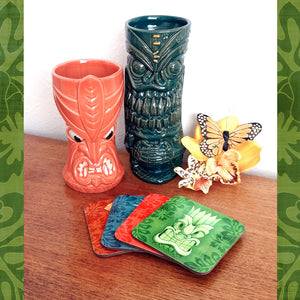 Set of four Tiki Coasters on a teak sideboard with two tiki mugs and some tropical flowers | The Inkabilly Emporium