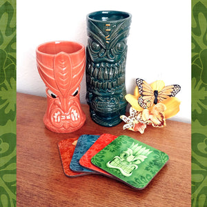 Set of four Tiki Coasters on a teak sideboard with two tiki mugs and some tropical flowers | The Inkabilly Emporium