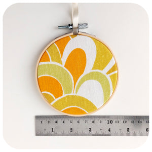 Vintage fabric framed in an embroidery hoop, featuring 70s swirl pattern in orange, yellow and green, next to a ruler to show scale | The Inkabilly Emporium