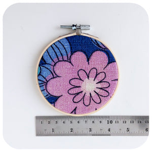A framed fabric hoop featuring a midcentury barkcloth floral design in lilac and blue, next to a ruler to show scale | The Inkabilly Emporium