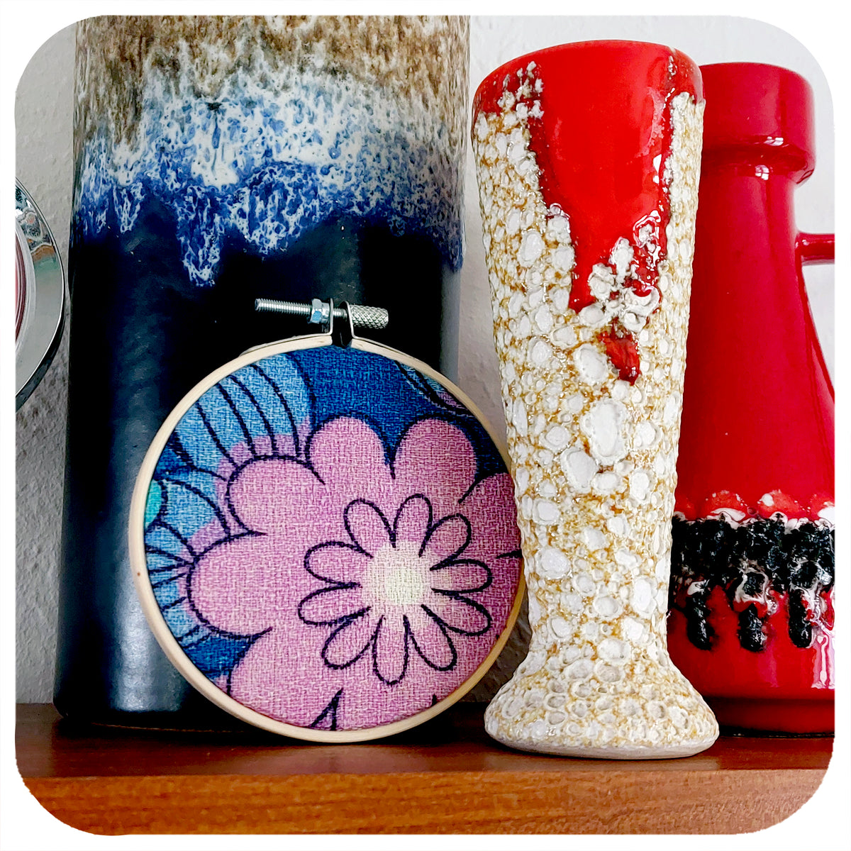 A framed fabric hoop featuring a midcentury barkcloth floral design in lilac and blue, sits on a shelf with vintage pottery | The Inkabilly Emporium