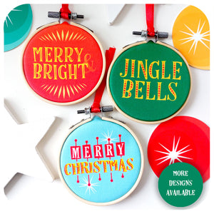 Three mid century style printed  fabric christmas decorations lie on a white background surrounded by white wooden stars and colourful discs. The text on the decorations reads: Merry & Bright, Jingle Bells and Merry Christmas.