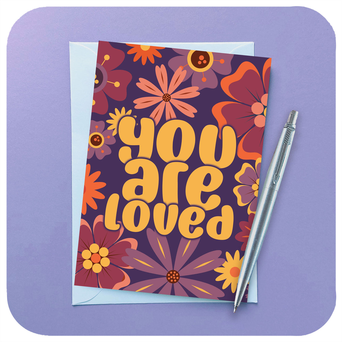 An A6 greetings card featuring 70s / 60s style flowers in pinks, purples & oranges and the words "You Are Loved" sits on a white envelope beside a chrome pen on a lilac background | The Inkabilly Emporium